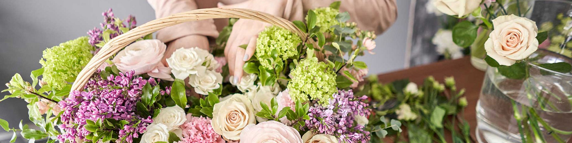 En person holder en kurv fyldt med farverige blomster, herunder roser, ranunkler og liljekonvaller. Der er også flere friske blomster på bordet, og stemningen er lys og indbydende.